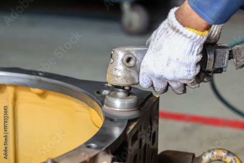 Tableau sur toile A technician uses a pneumatic orbital sander to polish the metal surface of a heavy yellow industrial component in a workshop, ensuring a smooth finish during a precision machinery overhaul