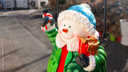 close up of a cute snowman figurine wearing green jacket and blue hat holding candy cane and red gift box for christmas decoration
