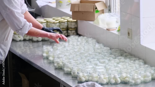 Gloved worker twists lids on glass jars filled with pickled vegetables on steel table. Worker stacks sealed jars in rows checking tight caps
