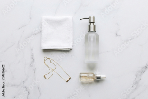 Minimalist bathroom accessories featuring a white towel glass dispenser bottle facial serum and a gold necklace on marble