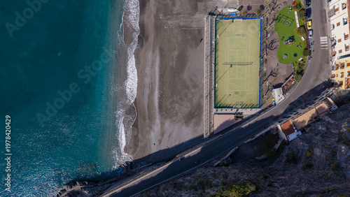 Aerial view of the dark sandy beach meeting the turquoise ocean next to a tennis court and colorful buildings, Minori, Salerno Italy