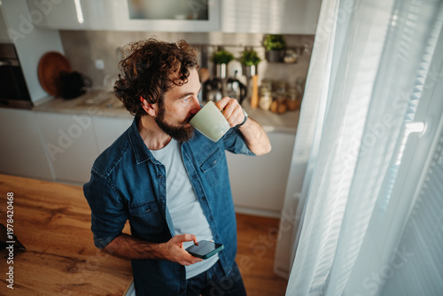 Man drinking coffee and using smartphone in kitchen