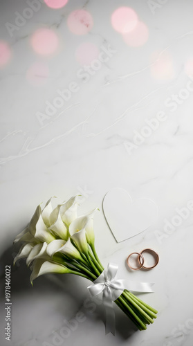 White calla lilies tied with a ribbon. Gold rings rest beside a heart-shaped cutout. Marble background with soft pink bokeh. Elegant wedding or engagement theme. Perfect for romantic, bridal