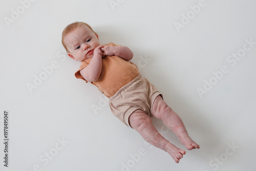 A full-length overhead shot of an infant lying on a white background. The baby wears a rust-colored ribbed top and beige shorts. High angle studio perspective