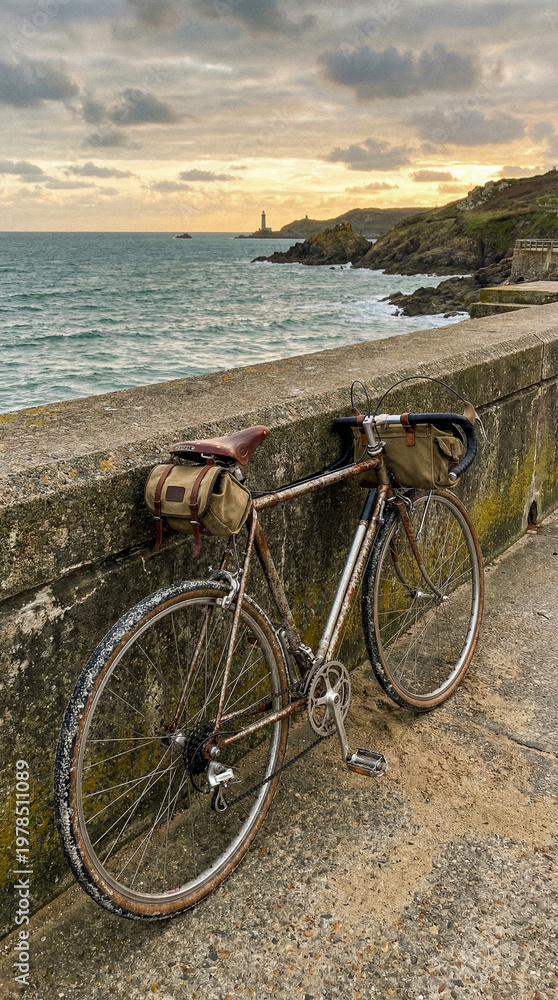Fototapeta premium Bicicleta clásica apoyada en un paseo marítimo durante un atardecer nublado. El mar picado, un faro lejano y acantilados completan esta escena melancólica y aventurera junto al océano.