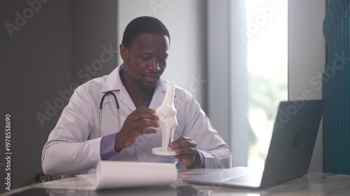 African American male physical therapist doctor looking at laptop holding anatomical knee joint model in rehabilitation center, Orthopedic medical clinic, healthcare and physiotherapy concept
