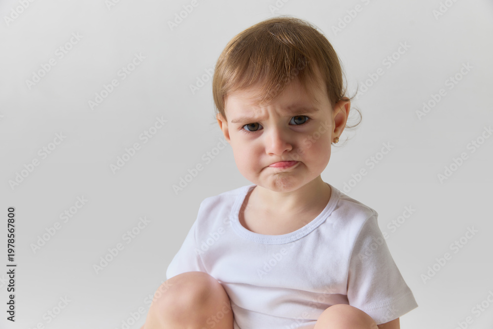 Naklejka premium Toddler girl sitting and making sad face with emotional expression isolated on white background. Concept of child emotions, mood changes, early psychology and communication of feelings.