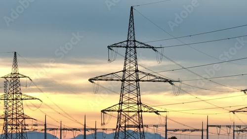 Power lines and electrical transmission towers stand tall in the rural area as the sun sets behind them. The sky shows vibrant colors.