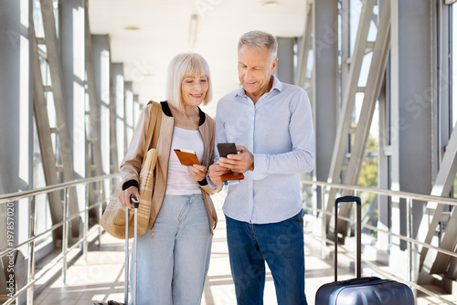 Mature couple standing in sunlit airport corridor, man using smartphone while woman holds passport and boarding pass, both smiling at screen, ready for travel