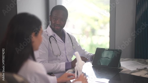 African American orthopedic doctor explaining knee joint x-ray on digital tablet to female colleague, Medical team consultation, bone anatomy model, diagnosis and healthcare technology concept
