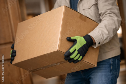 Person carrying heavy shipping carton indoors, green and black work gloves and casual jacket