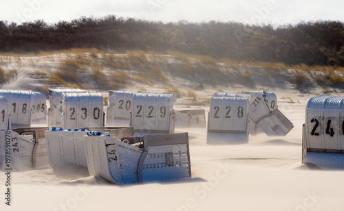 White beach chairs on the sandy beach of Spiekeroog, a North Sea island in Wadden Sea National Park (East Frisia, Lower Saxony, Germany). On a sunny stormy spring day, strong winds blow fine sand.
