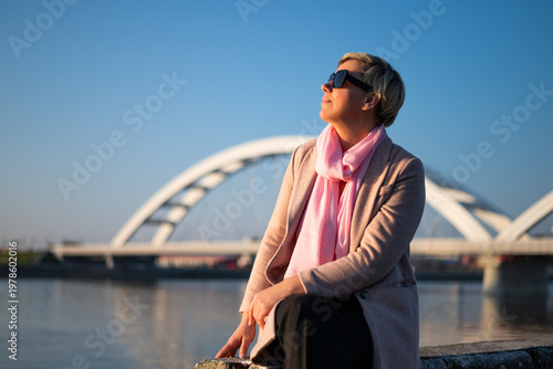 Portrait of urban woman with sunglasses sitting by the city river, enjoying sunshine, calm and thoughtful mood, happiness and freedom.