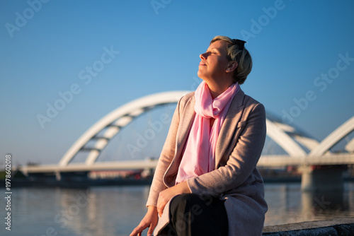 Portrait of beautiful natural woman sitting by the river in a city, enjoying sunshine, calm thoughts and a moment of pure happiness.