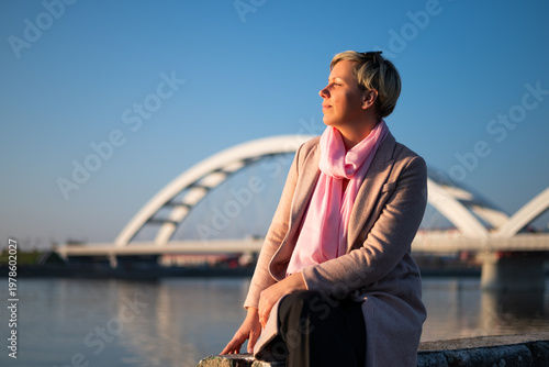 Portrait of a carefree woman sitting by the city river, looking away thoughtfully and enjoying quiet time on a sunny day.