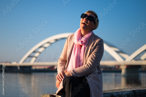 Portrait of smiling woman in sunglasses sitting by riverbank, enjoying freedom and calm, urban city background, sunlight reflection, carefree lifestyle moment.