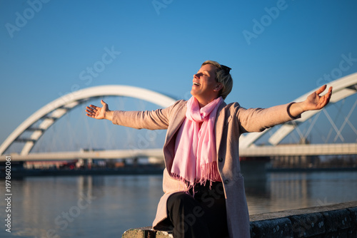 Portrait of cheerful woman with short blond hair smiling with arms raised on a sunny day, embracing freedom and happiness by the city river.