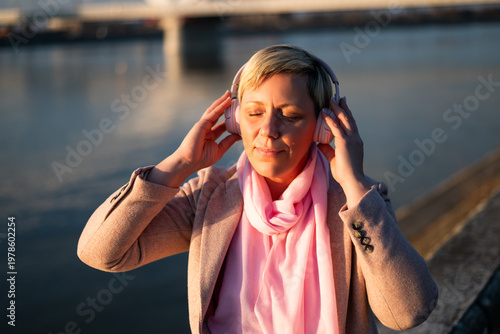 Portrait of relaxed woman with a short blonde hair listening music through wireless headphones and smiling while sitting by river in the city on a sunny day.	