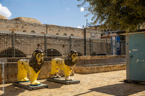 Jerusalem, Israel, Two whimsical yellow lion statues with , stand on stone pavement near a metal fence and historic buildings at Mahse Square, in the area of Beit Mahseh and Beit Rothschild.