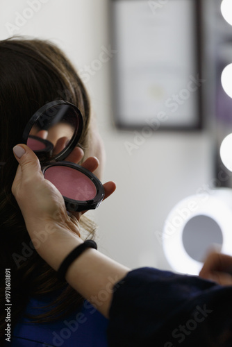 Makeup artist applying pink blush with brush on woman's cheek while she holds open compact mirror, professional beauty session in bright studio with ring light. Makeup artist work in beauty studio