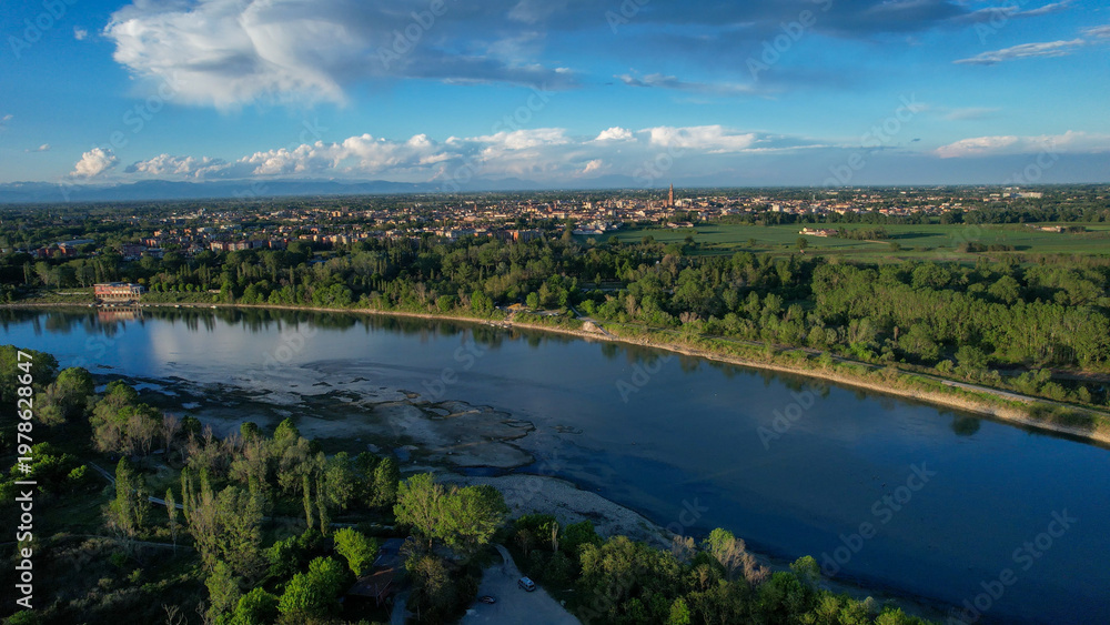 Fototapeta premium Po River marking the Emilia Romagna and Lombardy border with Cremona's skyline and Torrazzo tower rising above green floodplain fields under a summer sky