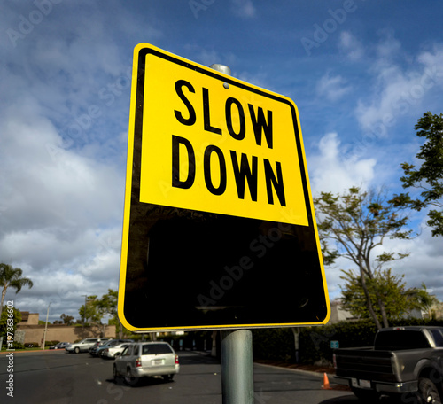 фотографія A yellow sign in a parking lot stating Slow Down against a blue sky