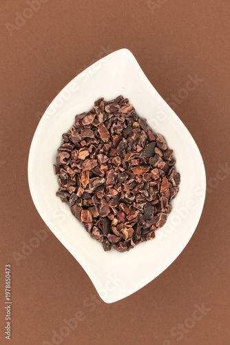 Cacao nibs in white leaf shaped bowl on brown background, overhead view