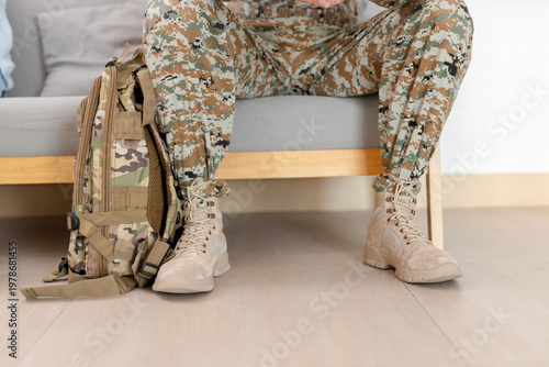 Military soldier legs in camouflage uniform with tactical boots and backpack sitting on sofa at home, army service concept, returning from duty, rest, transition to civilian life.