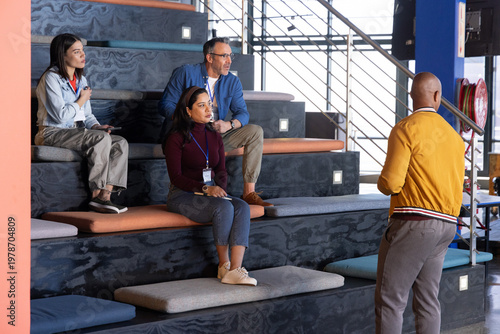 Diverse coworkers sitting on tiered benches in coworking space, wearing lanyards, facing presenter