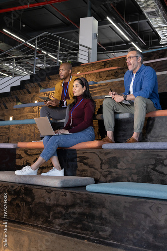 Diverse coworkers sitting on tiered wooden seating at conference using laptop, smartphone, lanyards