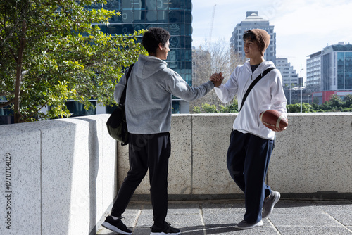 Clasping hands, holding football, diverse teenage males standing on rooftop with bags in sportswear