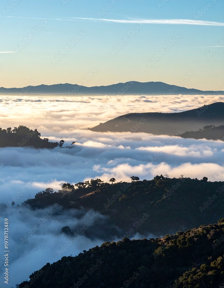 Fototapeta premium Aerial landscape of rolling hills emerging from a sea of clouds, with distant mountains under a blue sky. Sunlight illuminates the scene