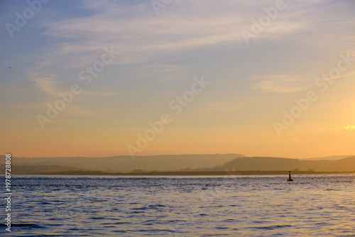 March 21 2026. A serene sunset seascape from Exmouth, a lone navigation buoy on the golden waters of the Exe estuary, hazy East Devon hills fading into the orange horizon.