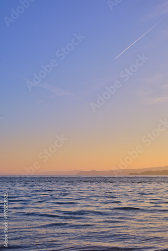 A minimalist dusk seascape from Exmouth with calm sea, a golden-to-blue gradient sky and a solitary aeroplane contrail streaking across the Devon coastline.