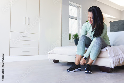 Foto Asian woman sitting on low white bed edge tying black sneakers in bedroom, smart