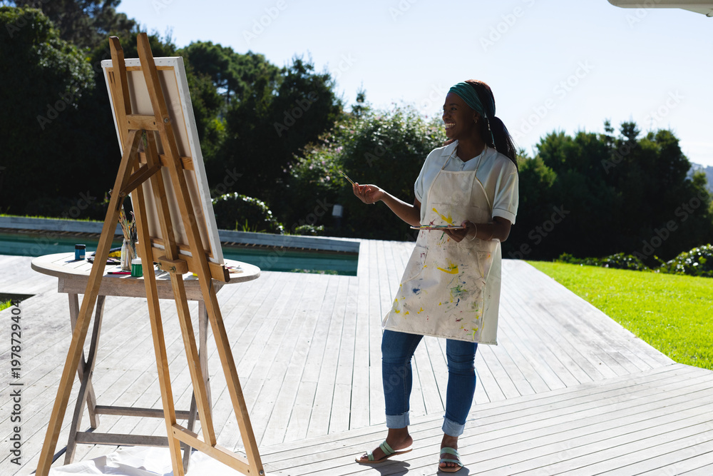 Naklejka premium Mid-adult African American woman painting at easel on deck beside pool wearing apron, copy space