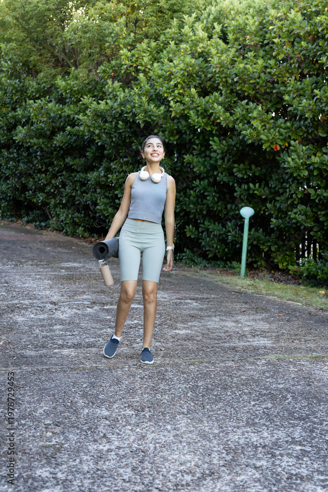 Fototapeta premium Indian woman walking on paved path by hedge in activewear carrying yoga mat, bottle, headphones