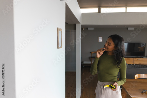 Woman holding tape measure and pencil measuring picture placement on white wall at home, copy space