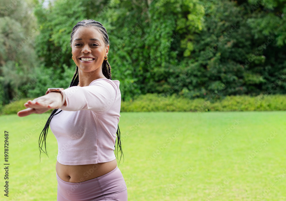 Fototapeta premium Mid-adult African-American woman reaching arm on lawn, wearing pink set, tracker, hoops, copy space