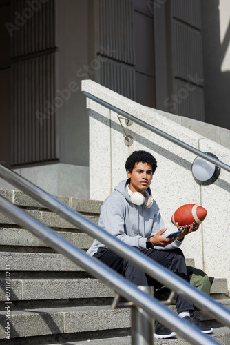 African American man sitting on campus steps holding football smartphone and headphones