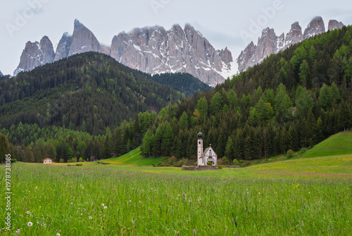 Scenic alpine meadow with a small chapel nestled among lush green forests and dramatic snow-dusted mountain peaks under a misty sky, evoking tranquility and natural beauty.