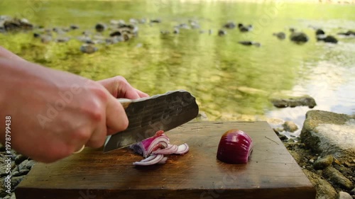 Close up of chopping onion near beautiful clean river at calm summer day. Young man cutting purple onion with sharp knife on wooden board. Cooking outdoor concept
