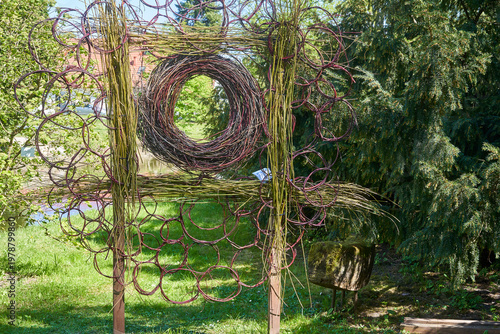 A large outdoor sculpture made of woven willow branches and twigs stands in a lush green botanical garden. The installation features two vertical wooden posts supporting horizontal woven elements,