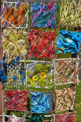 A vibrant outdoor art installation composed of various natural and recycled materials arranged in a grid pattern within square sections. The artwork features an eclectic mix of dried plants, painted