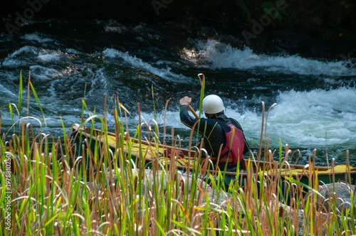 Valokuva A lone canoer paddles upstream on fast river rapids near marshy reeds while wearing a helmet and life jacket