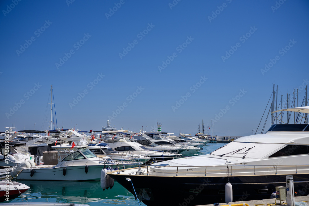 Fototapeta premium Luxury yachts, sailboats moored at sunny Mediterranean marina. Nautical lifestyle, high-end travel and summer vacation concept, marine transportation. Marina harbor with turquoise water under sky