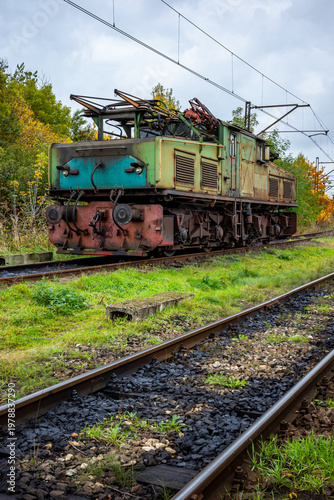 An old, green, electric shunting locomotive standing on a side track. The photo was taken on a cloudy autumn day.