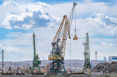 Принти на полотні Industrial harbor scene featuring massive portal cranes against a cloudy sky, with a scrap metal pile and coastal hills in the background