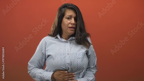 Woman middle aged brunette in striped shirt with hand on lower stomach and pained expression in studio with red wall; pain concern.