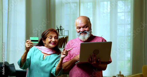 Senior Indian couple standing in a modern living room, using a debit card for an online transaction and showing it to the camera with confidence and smiles